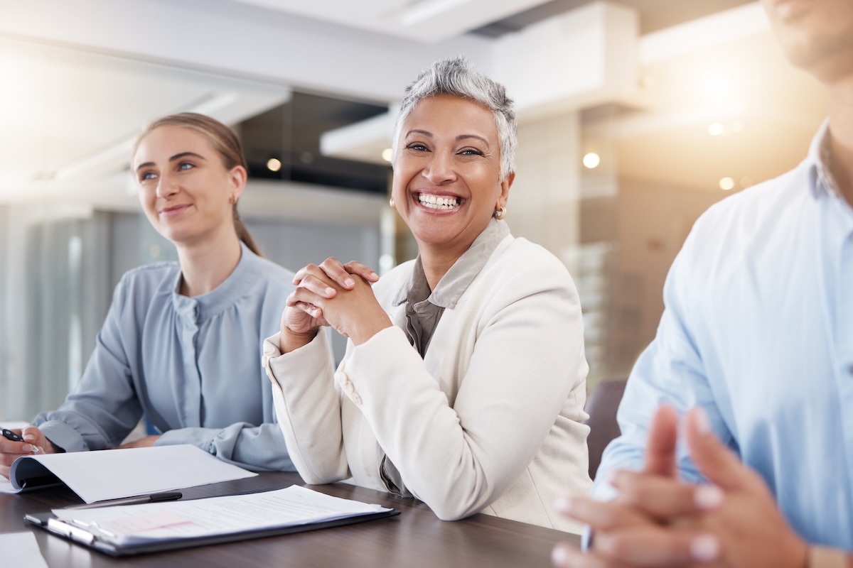 a woman with short grey hair sits in a corporate setting with two other people and smiles at the came